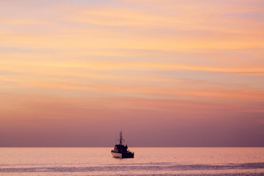 Wide shot of a professional offshore fishing boat heading out to the deep sea as the sun rises on the horizon, casting a g...