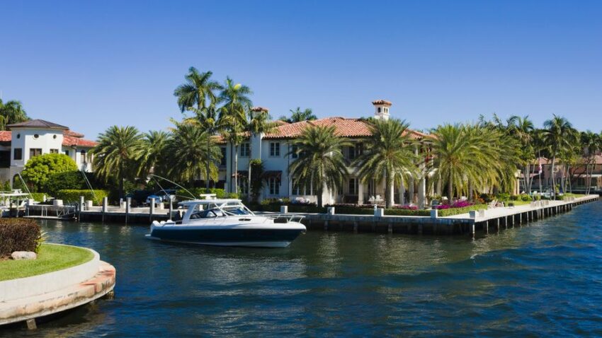 Bright, modern waterfront home with large windows and a private wooden dock where a boat is moored on a sunny day.